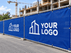 A blue construction hoarding banner with a white logo template reading 'YOUR LOGO' is attached to a chain-link fence at an outdoor construction site with a building and crane in the background.